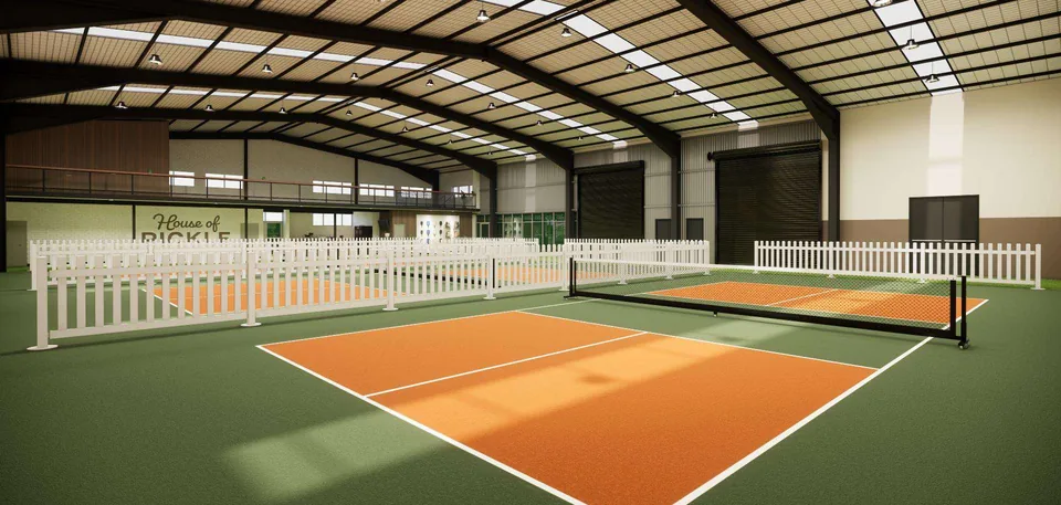 A large indoor hall with an orange and green pickleball court, white picket barriers and a mezzanine